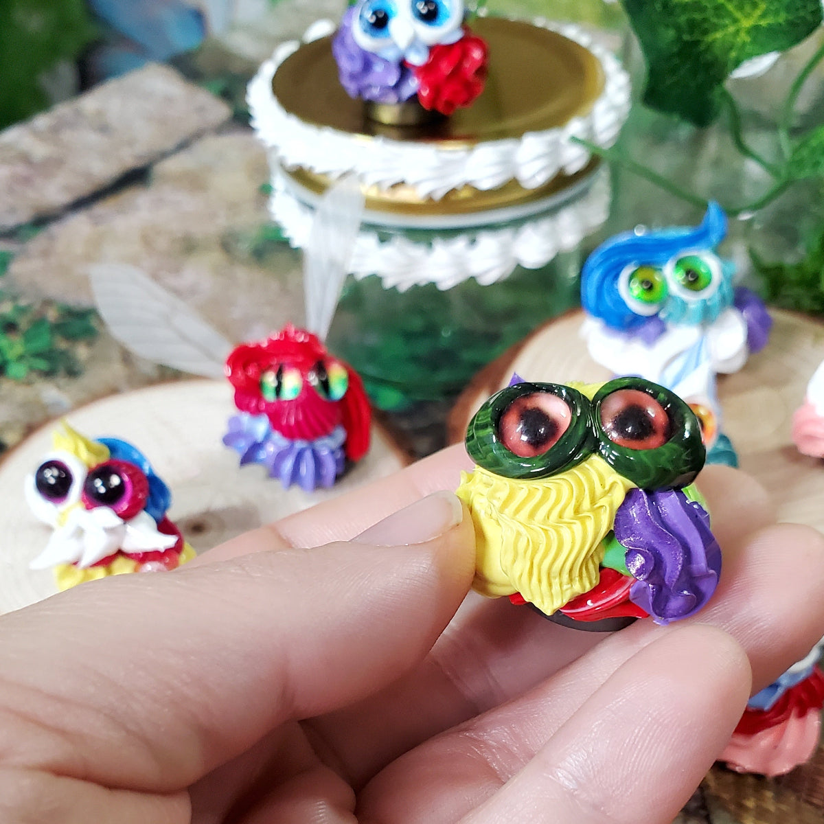 Hand holding a tiny green, yellow, red and purple Squirklee with various other Squirklees in the background.