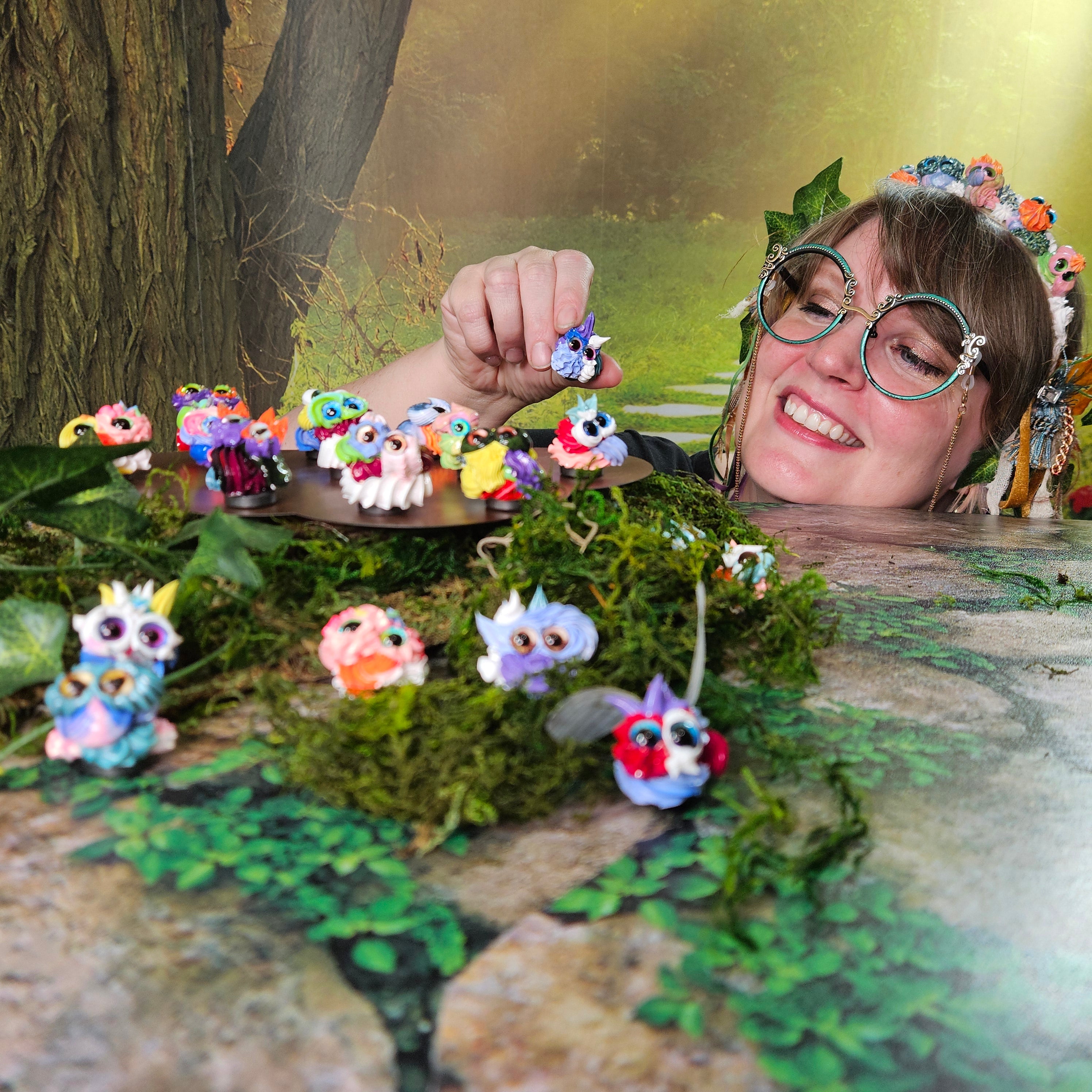 Person with frosting creature headband and big green glasses against a natural background, holding a tiny frosting creature called a squirklee