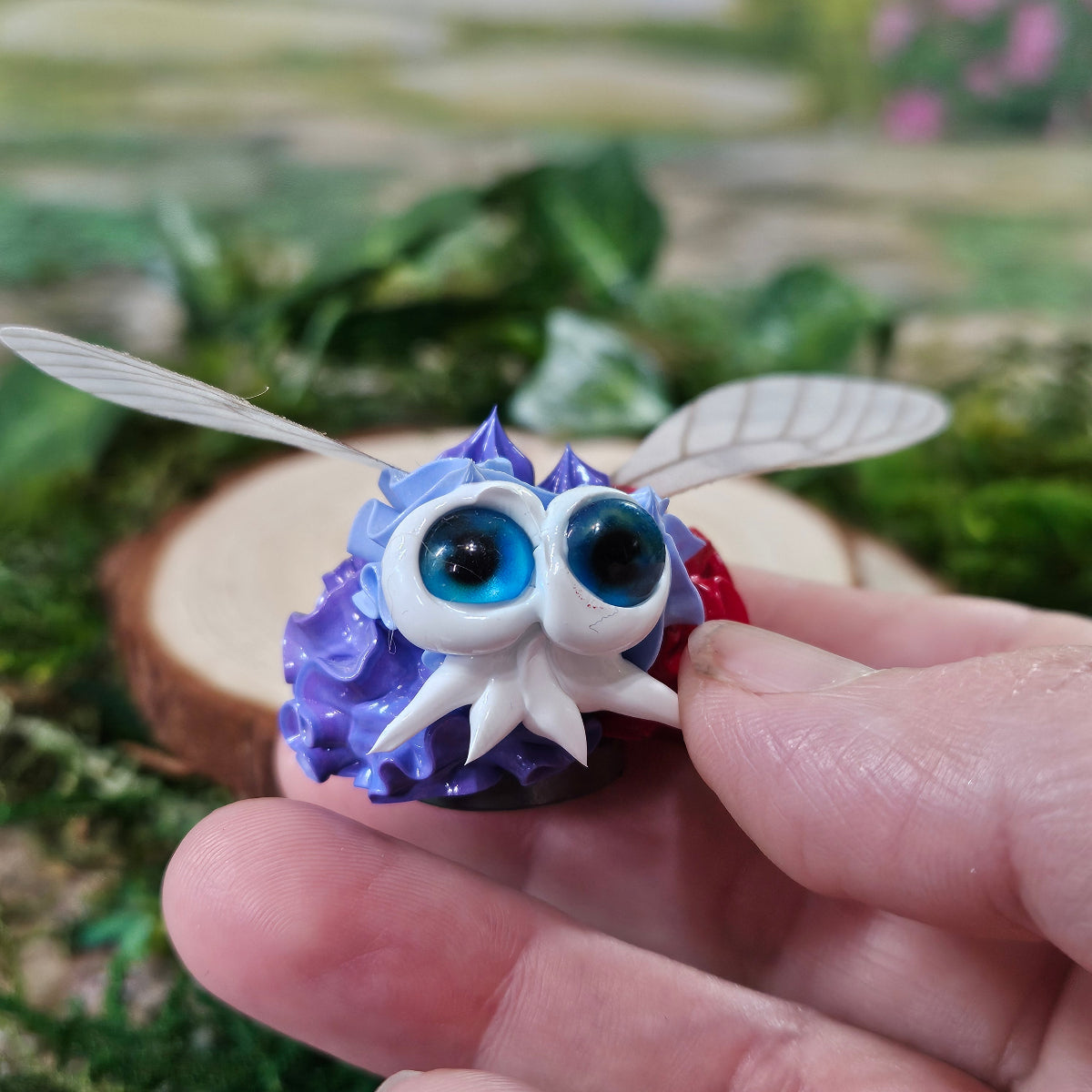Hand holding a purple, white and red Velvet Flutterling Squirklee. The Squirklee has wings and blue eyes.