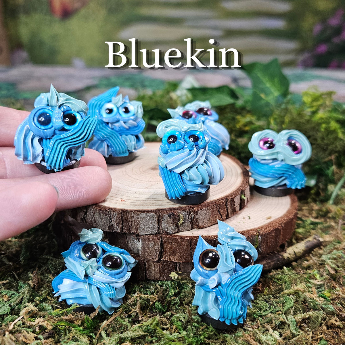 Six small blue frosting figurines with large  eyes are displayed on a rustic wood slice and mossy surface. A human hand is visible holding one on the left, and the word 'Bluekin' appears at the top.