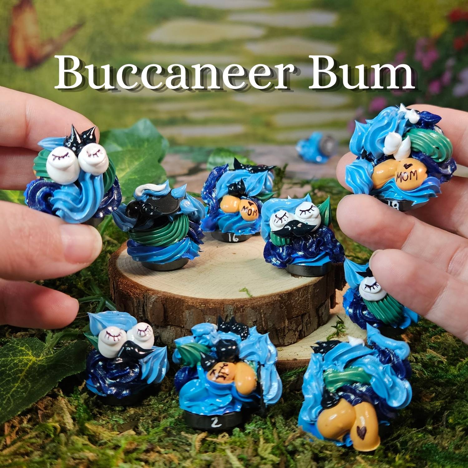 Several small blue and black fantasy frosting figurines are arranged on moss and a wood slice, with human hands holding some of them. Large text at the top reads 'Buccaneer Bum.