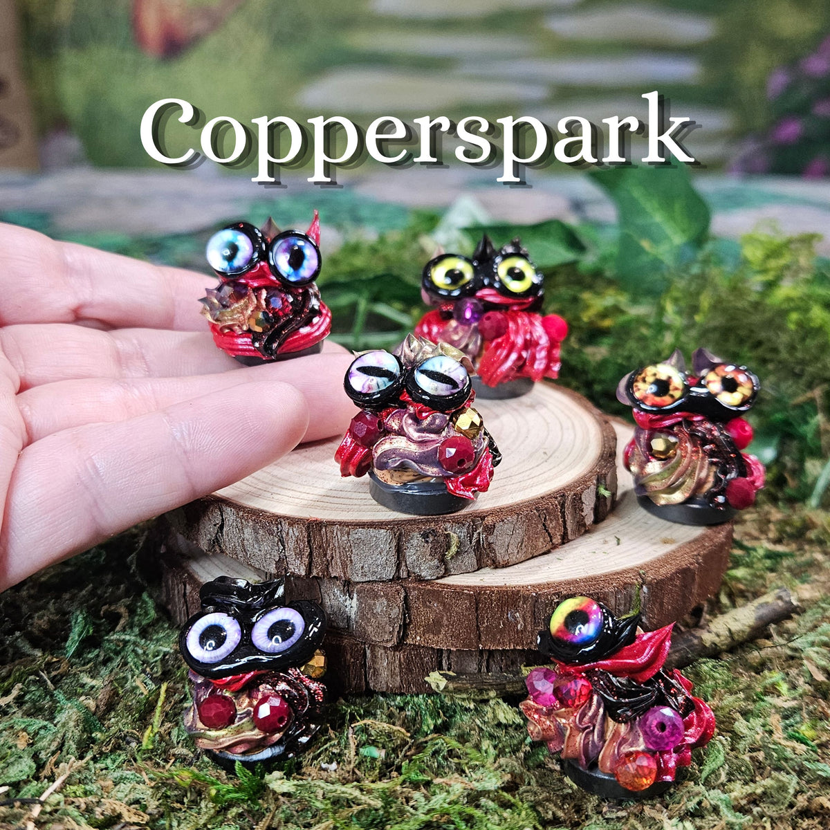 Five small colorful fantasy frosting figurines displayed on a mossy surface with a wood slice prop, plus a human hand holding one creature. Large text at the top reads 'Copperspark'.