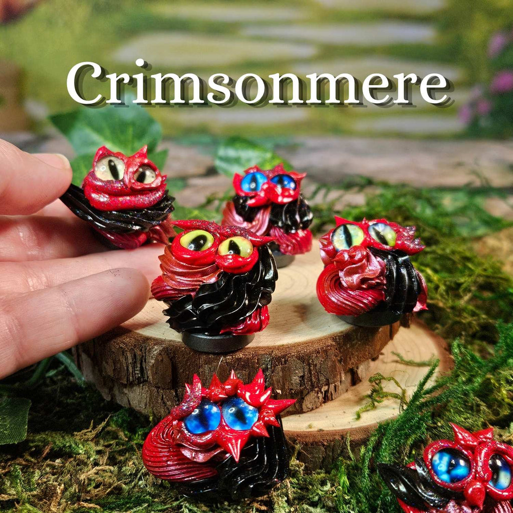 Several small frosting creatures are displayed on a wood slice with moss and a blurred outdoor-style background. A human hand is visible holding one of the creatures