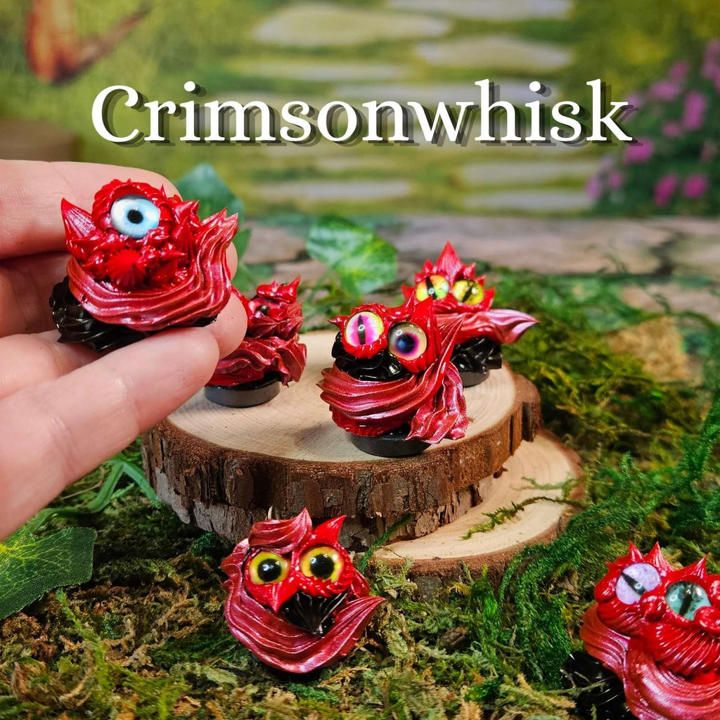 A hand holds a small red monster-shaped frosting creature with a large googly eye, while several similar frosting creatures are arranged on moss and wood slices in a miniature garden setting. The text 'Crimsonwhisk' appears at the top.