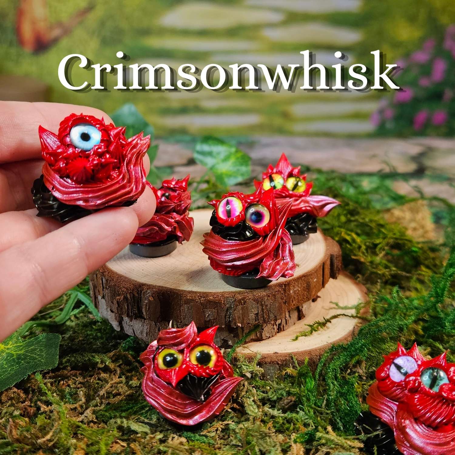 A hand holds a small red monster-shaped frosting creature with a large googly eye, while several similar frosting creatures are arranged on moss and wood slices in a miniature garden setting. The text 'Crimsonwhisk' appears at the top.