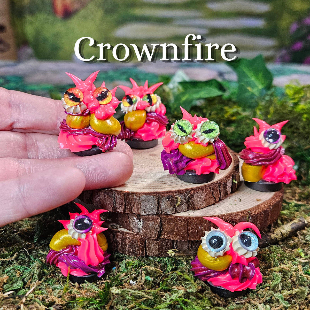 Five small fantasy creature figurines with pink wings and yellow bellies are displayed on moss and a wood slice, with a human hand holding one figure. The text 'Crownfire' appears at the top.