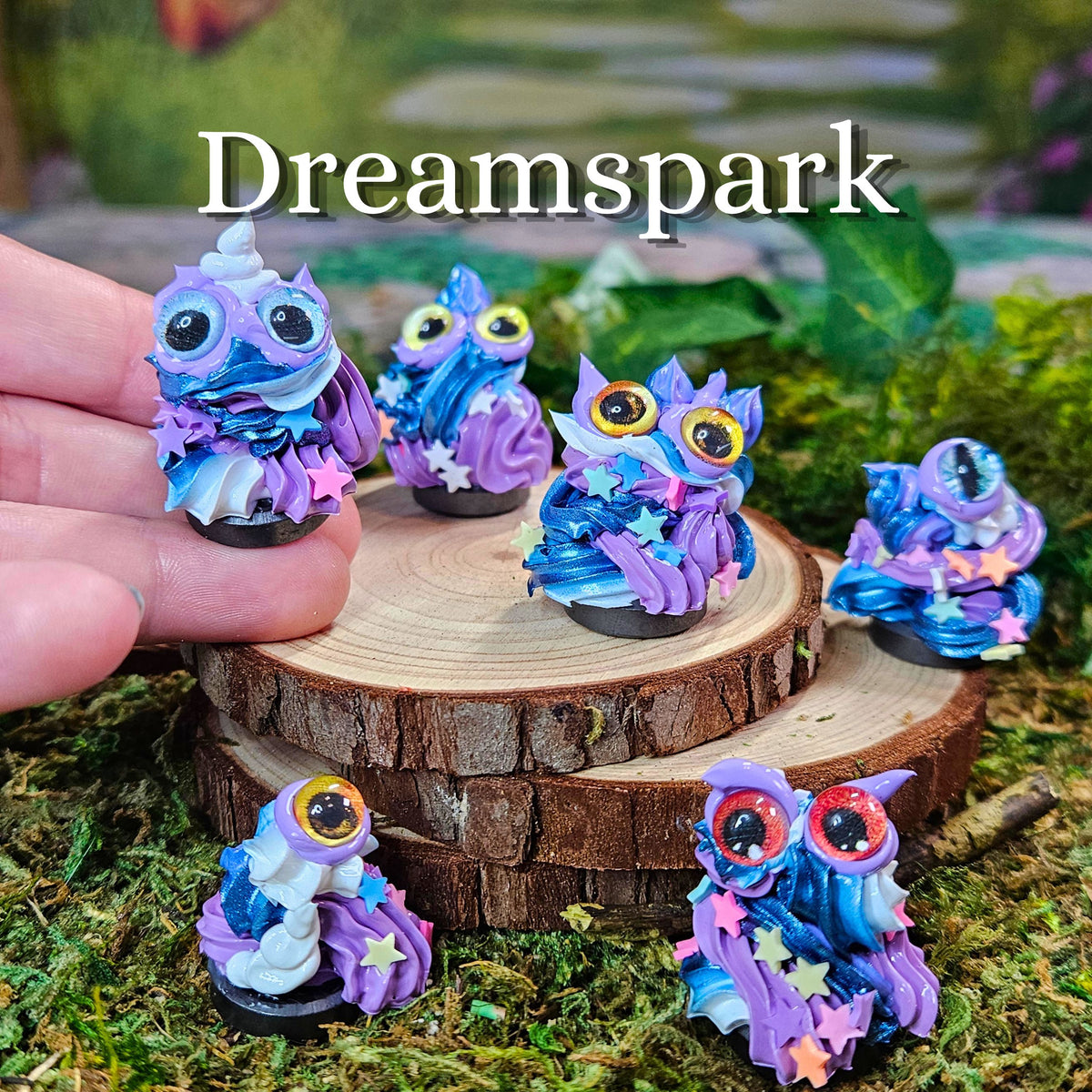 Five small fantasy frosting figurines in swirling blue, purple, and pink colors are displayed on a wood slice and mossy surface, with a human hand holding one on the left. The word 'Dreamspark' appears at the top.