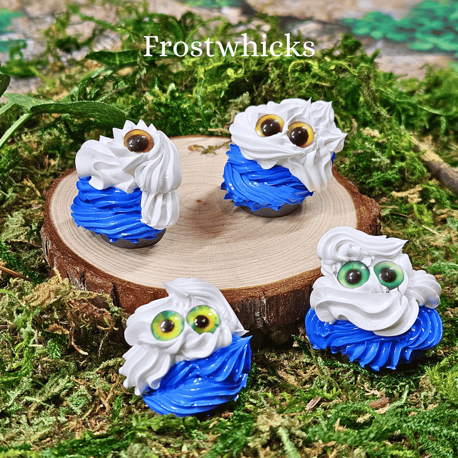 Four small blue and white frosting creatures on a wooden disc surrounded by green foliage.