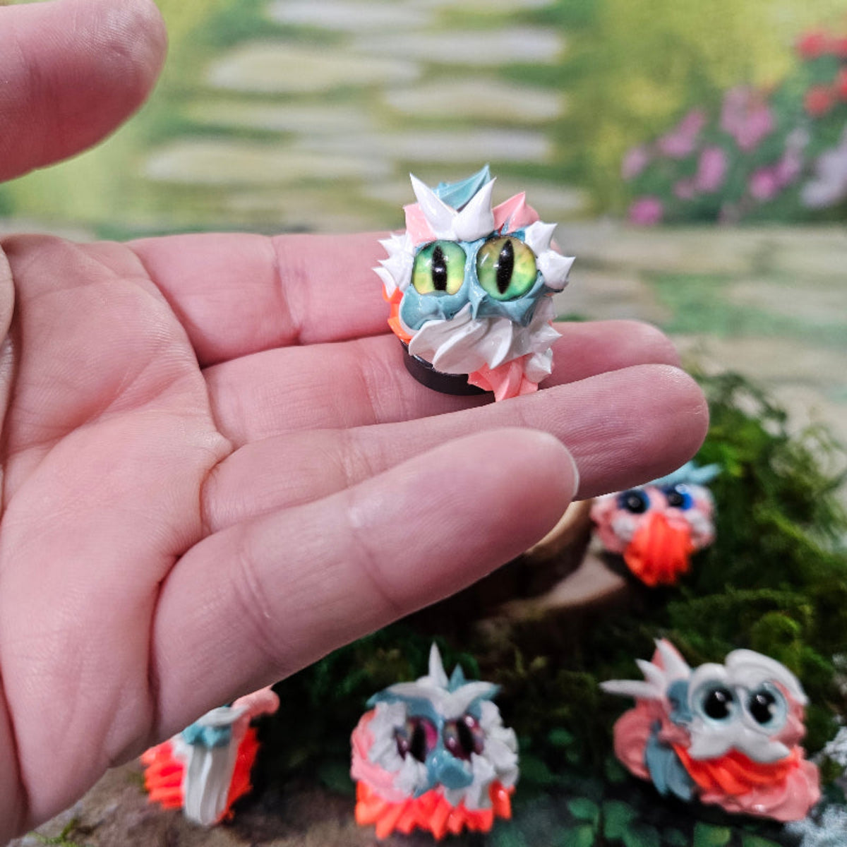 Hand holding a hand-made frosting creature called a Squirklee. The Squirklee has striking cat eyes, white , peach, orange and aqua "frosting".