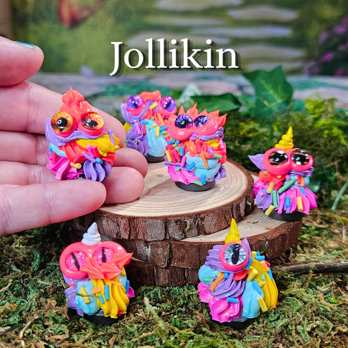 A hand holds a small colorful fantasy creature figurine labeled 'Jollikin' next to several matching figurines displayed on a wood slice and mossy surface. The creatures have bright pink, purple, blue, yellow, and orange colors with large glossy eyes.