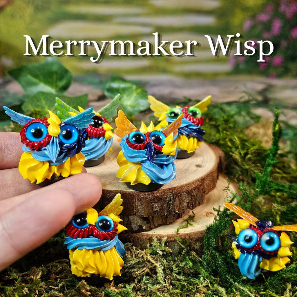 Five colorful handmade frosting figurines labeled 'Merrymaker Wisp' are displayed on moss and a wood slice, with a human hand holding one in the foreground. The creatures have blue and yellow bodies, colorful beaks, and large eyes