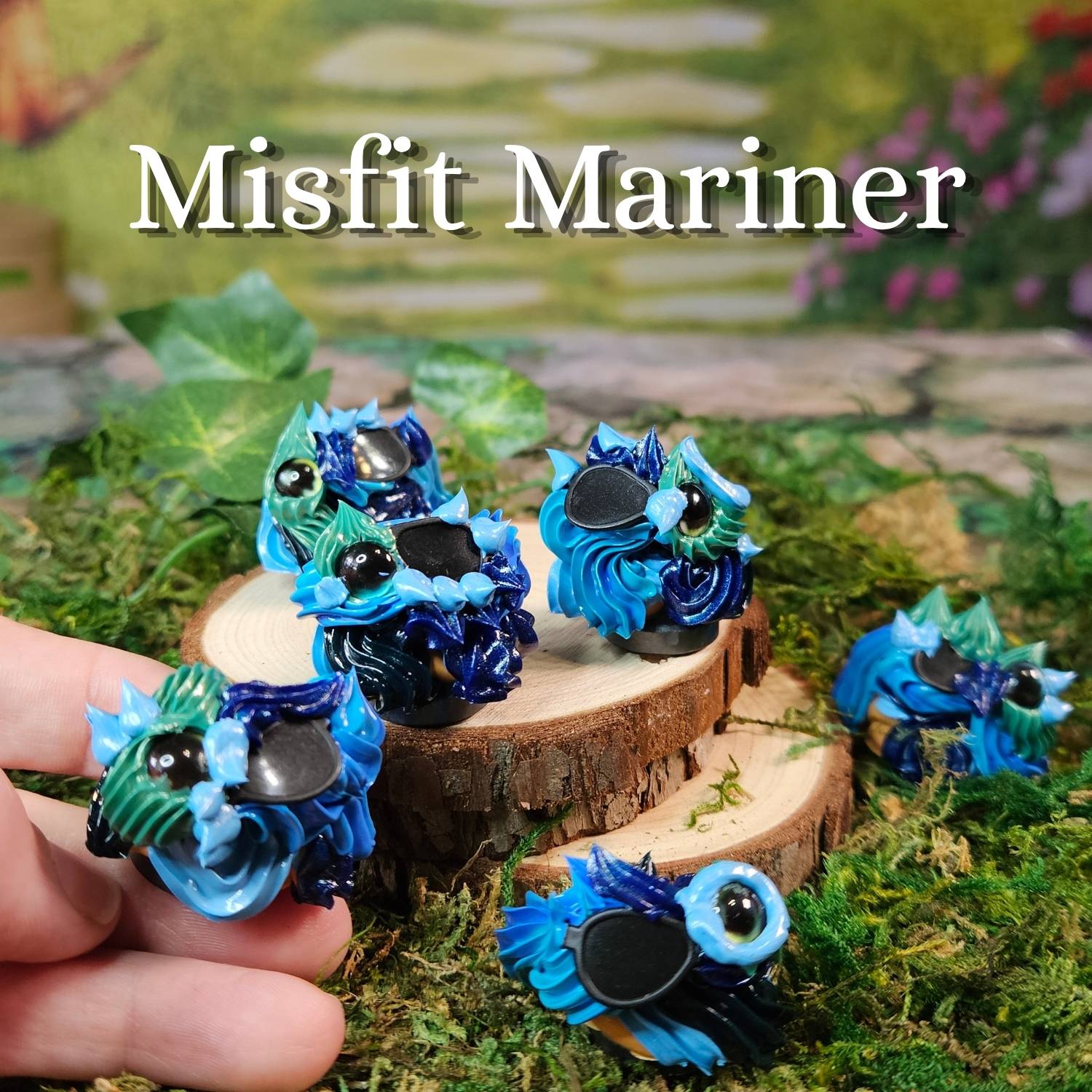 Several small decorative frosting figurines in bright blue, teal, and dark navy are displayed on moss and wood slices, with one held in a human hand. Large text at the top reads 'Misfit Mariner'.