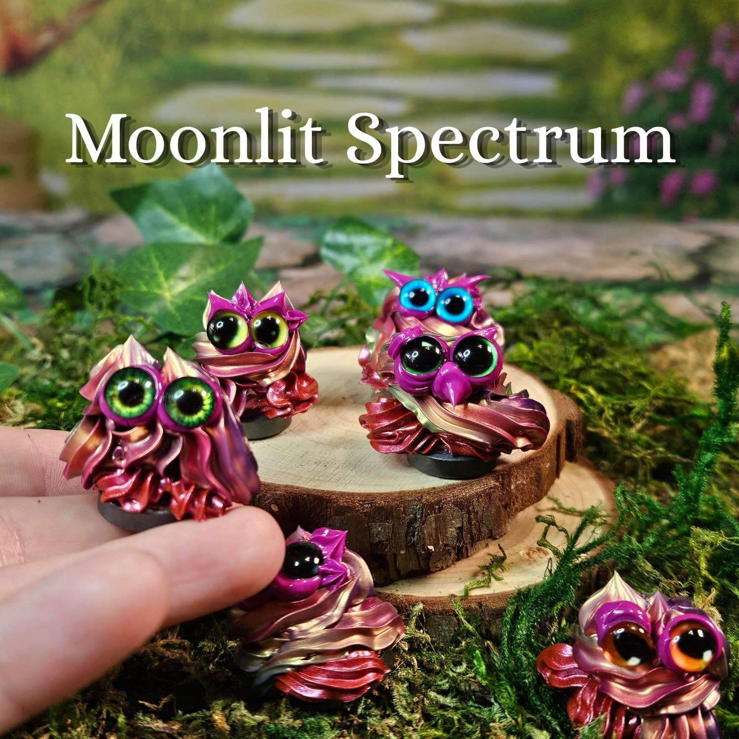 Four miniature frosting figurines with colorful iridescent sheen and large expressive eyes are displayed on moss, wood slices, and a hand, with the text 'Moonlit Spectrum' overlaid at the top.