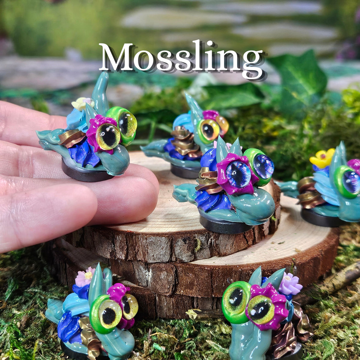 Several small frosting snail figurines are displayed on a mossy surface and a wooden slice, with a human hand holding one. The word 'Mossling' appears at the top.