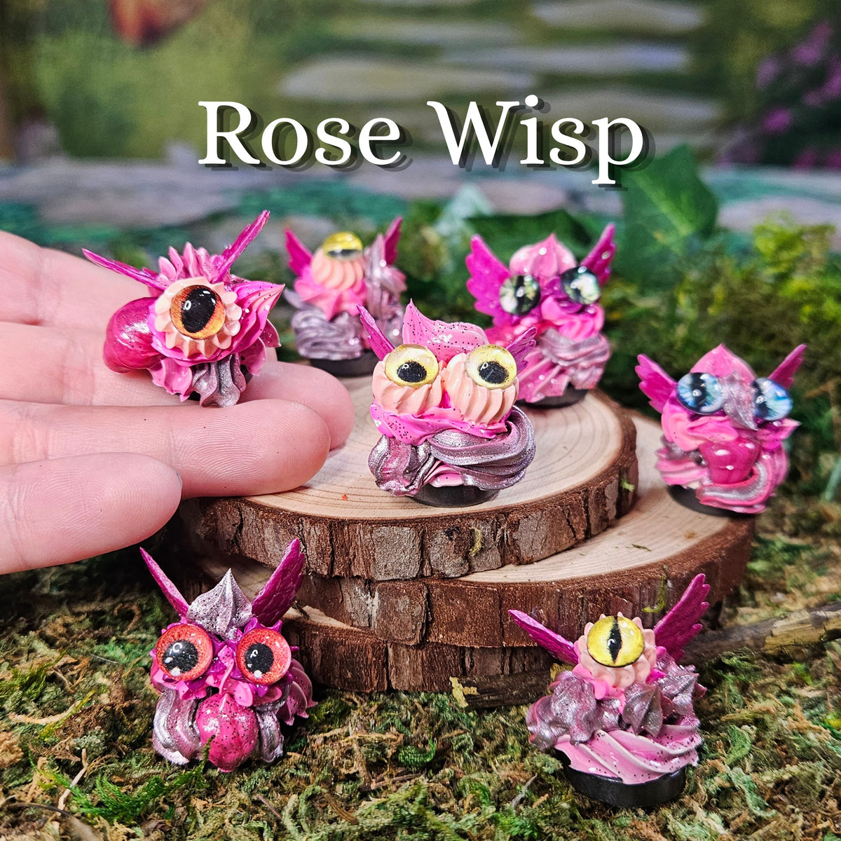 Several small pink fantasy frosting figurines are displayed outdoors on moss and wood slices, with one figurine being held in a human hand. The text 'Rose Wisp' appears at the top.