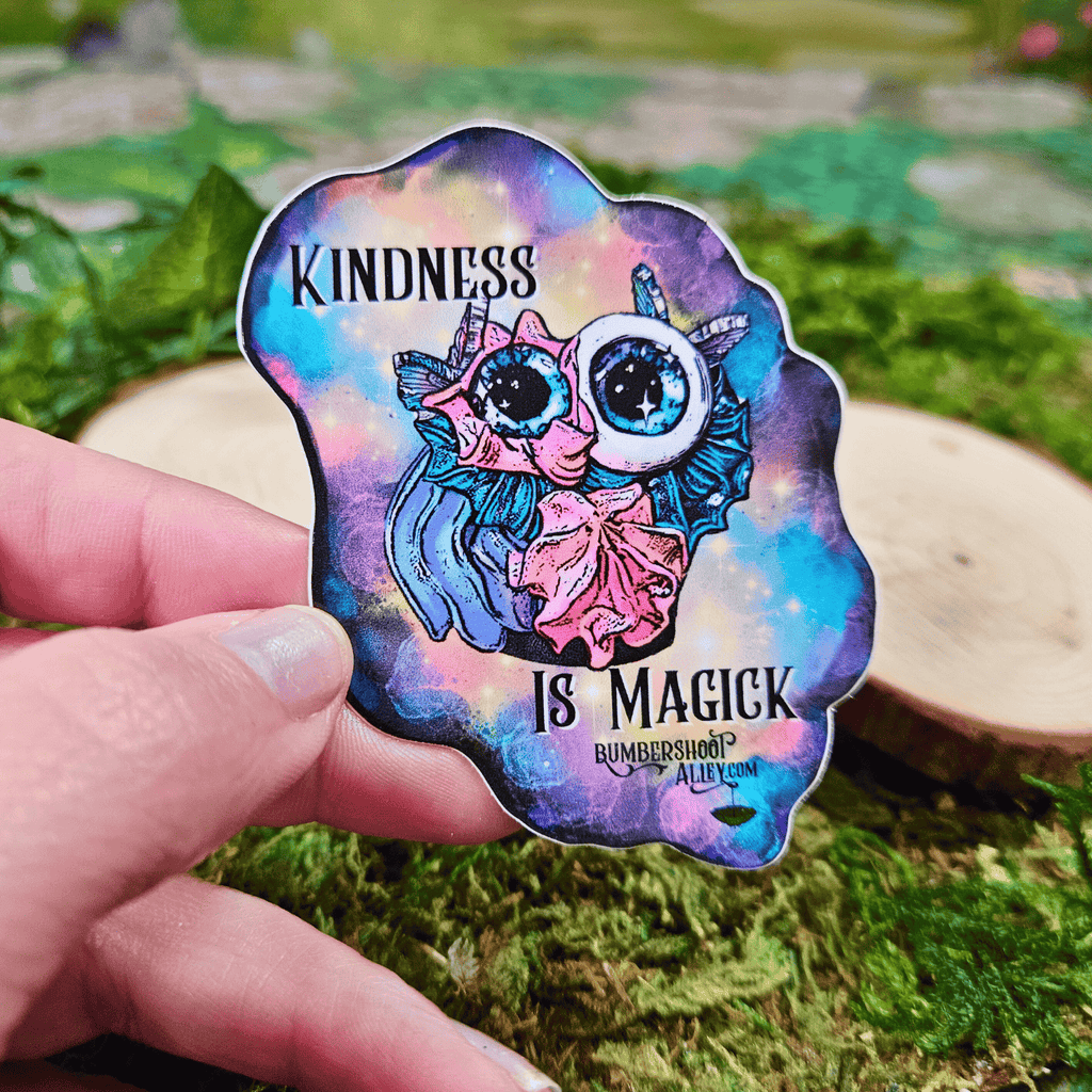 Colorful sticker with frosting creature and 'Kindness is Magick' text held by a hand on a natural background.