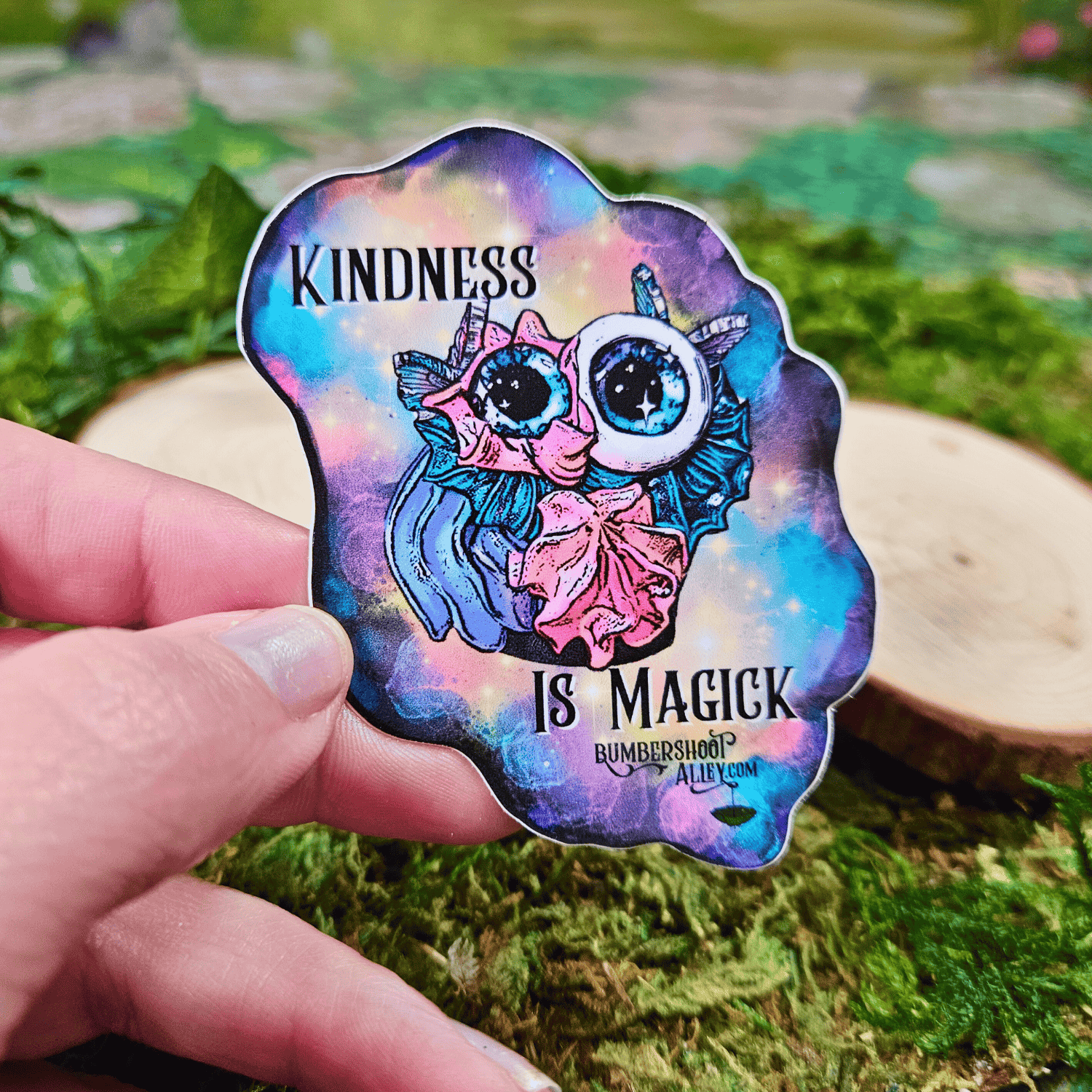 Colorful sticker with frosting creature and 'Kindness is Magick' text held by a hand on a natural background.