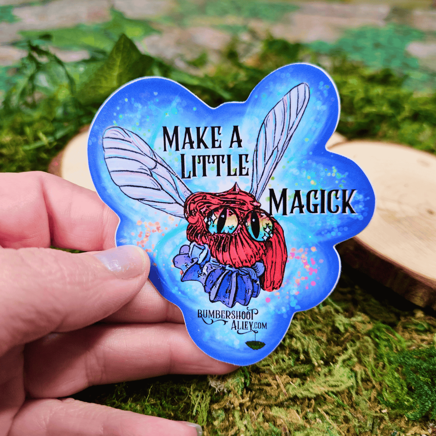 Sticker with a red character and text 'Make a little Magick' held by a hand on a natural background.