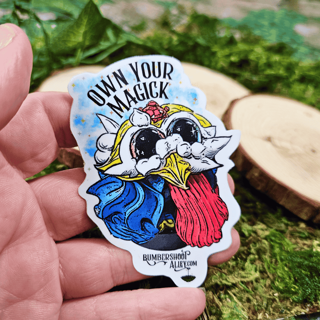 Sticker with 'Own Your Magick' text held by a hand against a natural background