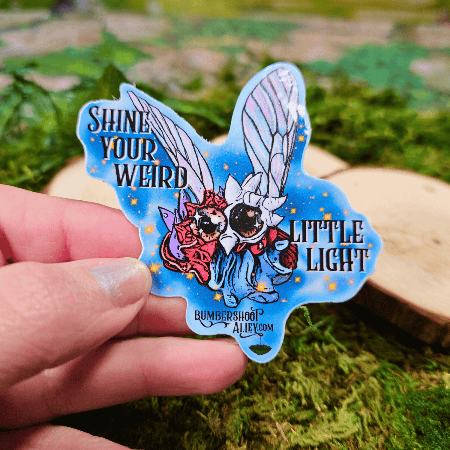 Sticker with winged frosting creature and text held by a hand on a natural background
