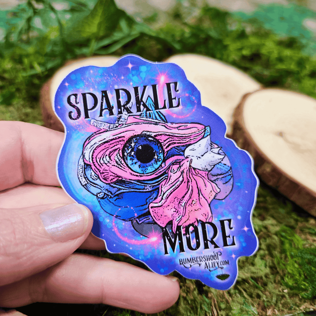 Sticker with one-eyed frosting creature and 'Sparkle More' text held by a hand on a natural background