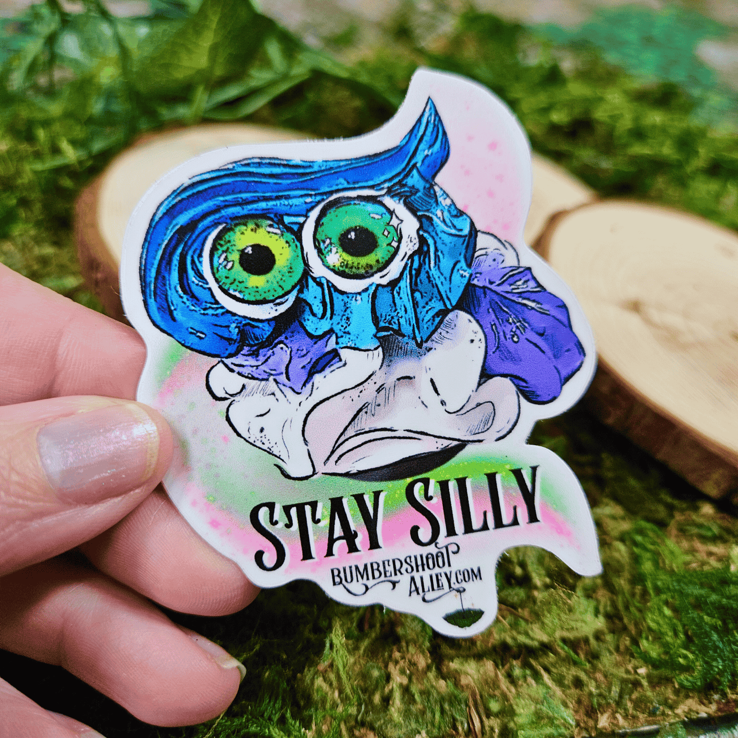 Sticker with a colorful frosting creature design held by a hand on a green leafy background