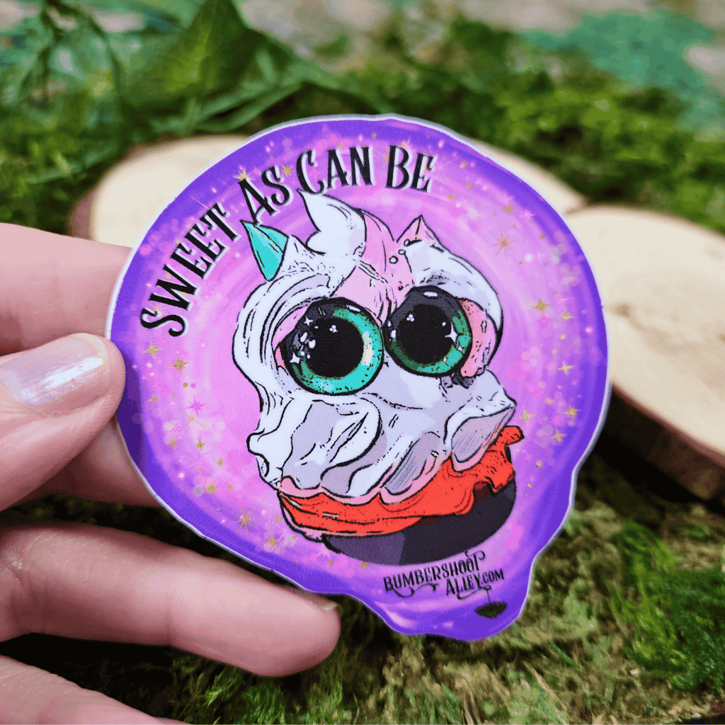 Sticker with a frosting creature and 'Sweet as can be' text held by a hand on a natural background