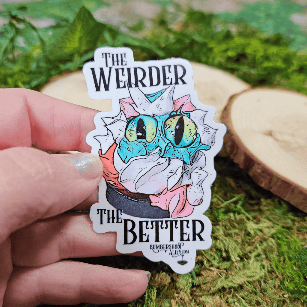 Sticker with frosting creature and text 'The Weirder The Better' held by a hand on a green leafy background.