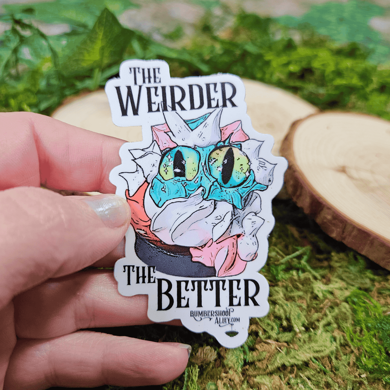 Sticker with frosting creature and text 'The Weirder The Better' held by a hand on a green leafy background.
