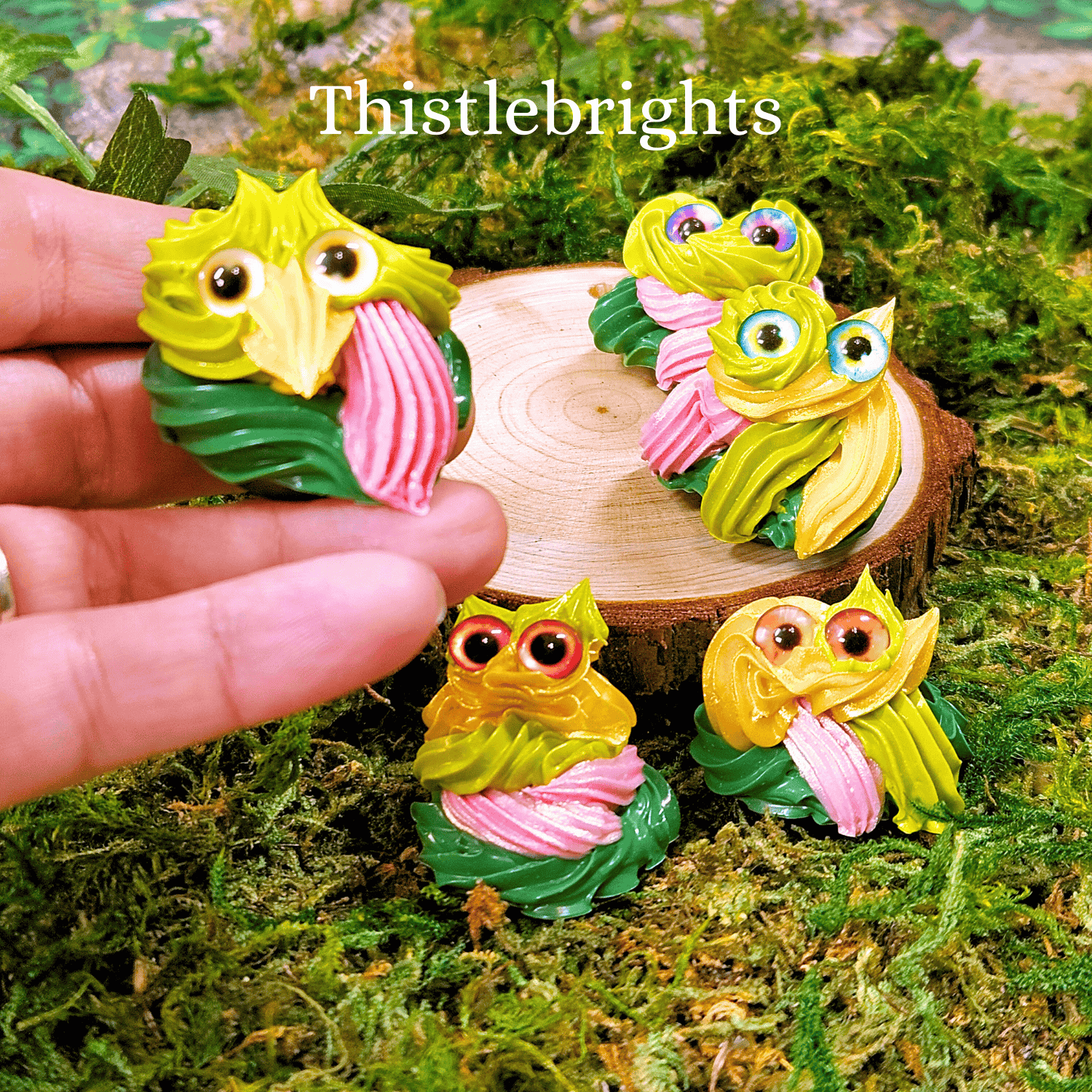Colorful frosting creatures on a wooden stump with 'Thistlebrights' branding, surrounded by greenery.