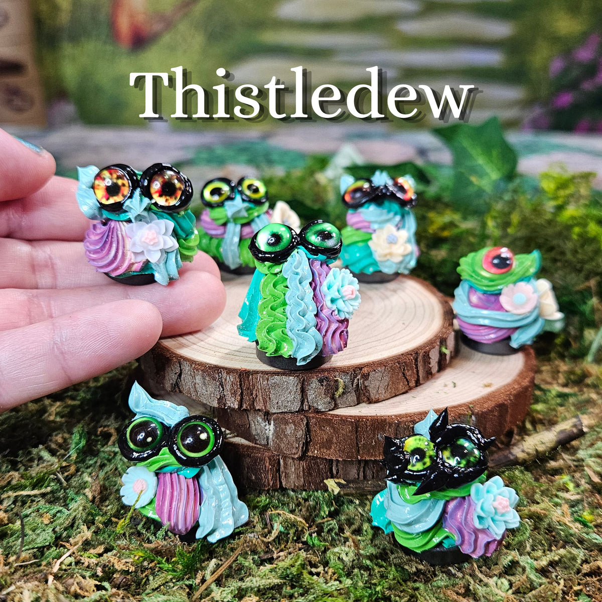 Several small frosting figurines are displayed on and around a wooden slice stand, with a human hand holding one piece on the left. The background is a blurred outdoor mossy setting.