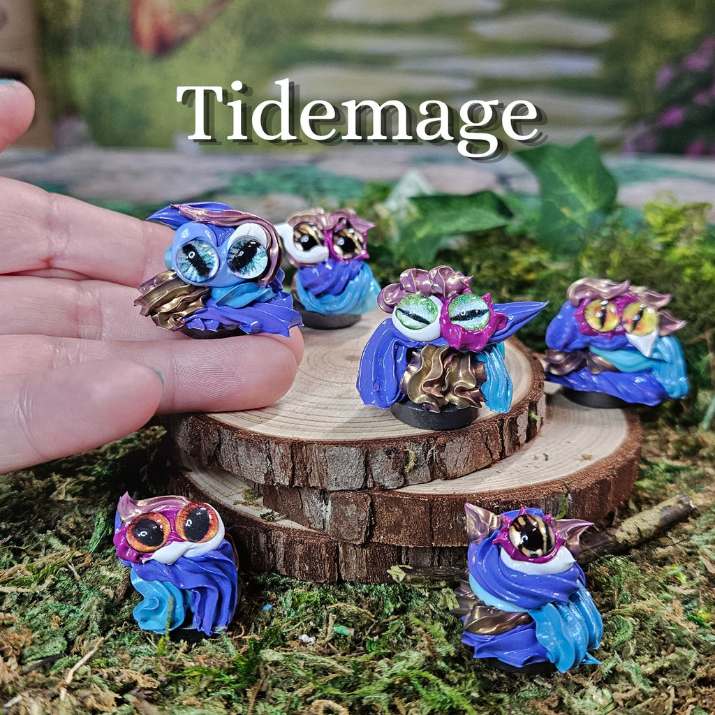 Five small frosting figurines with large eyes and colorful accents are displayed on moss and a wood slice, with a human hand holding one owl. The text 'Tidemage' appears at the top.