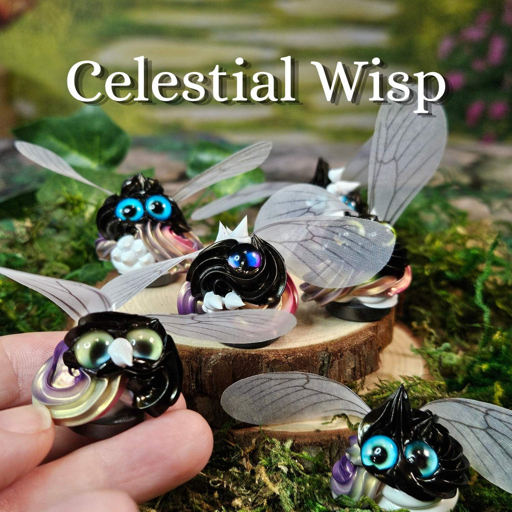 Several small fantasy frosting figurines with translucent wings, glossy black bodies, bright eyes, and pastel accents are displayed on moss and a wood slice. A human hand holds one figurine in the lower left, and the words 'Celestial Wisp' appear across the top.
