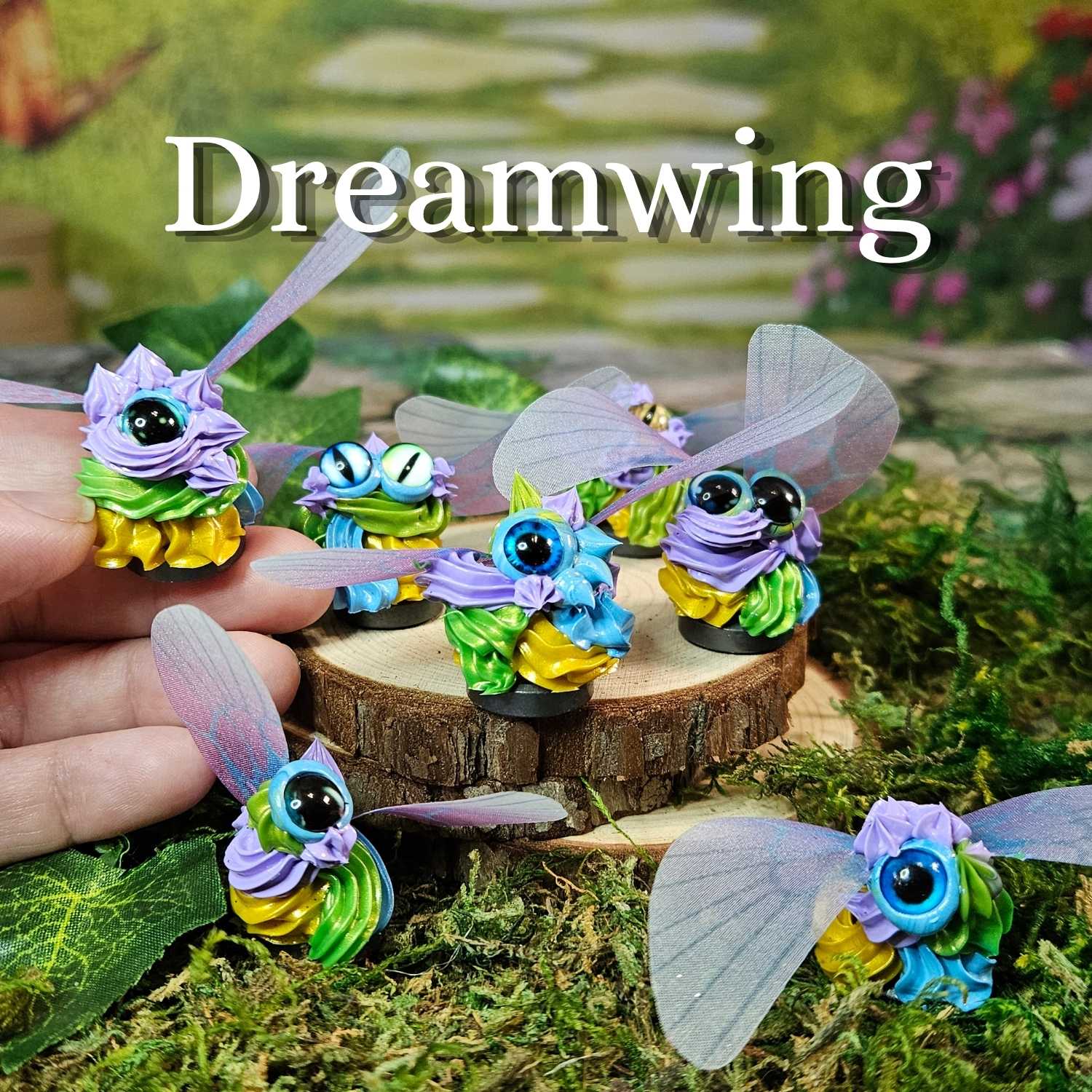 Five colorful fantasy frosting figurines with translucent wings are displayed on moss and a small wood slice, with a human hand holding one on the left. The word 'Dreamwing' appears at the top.