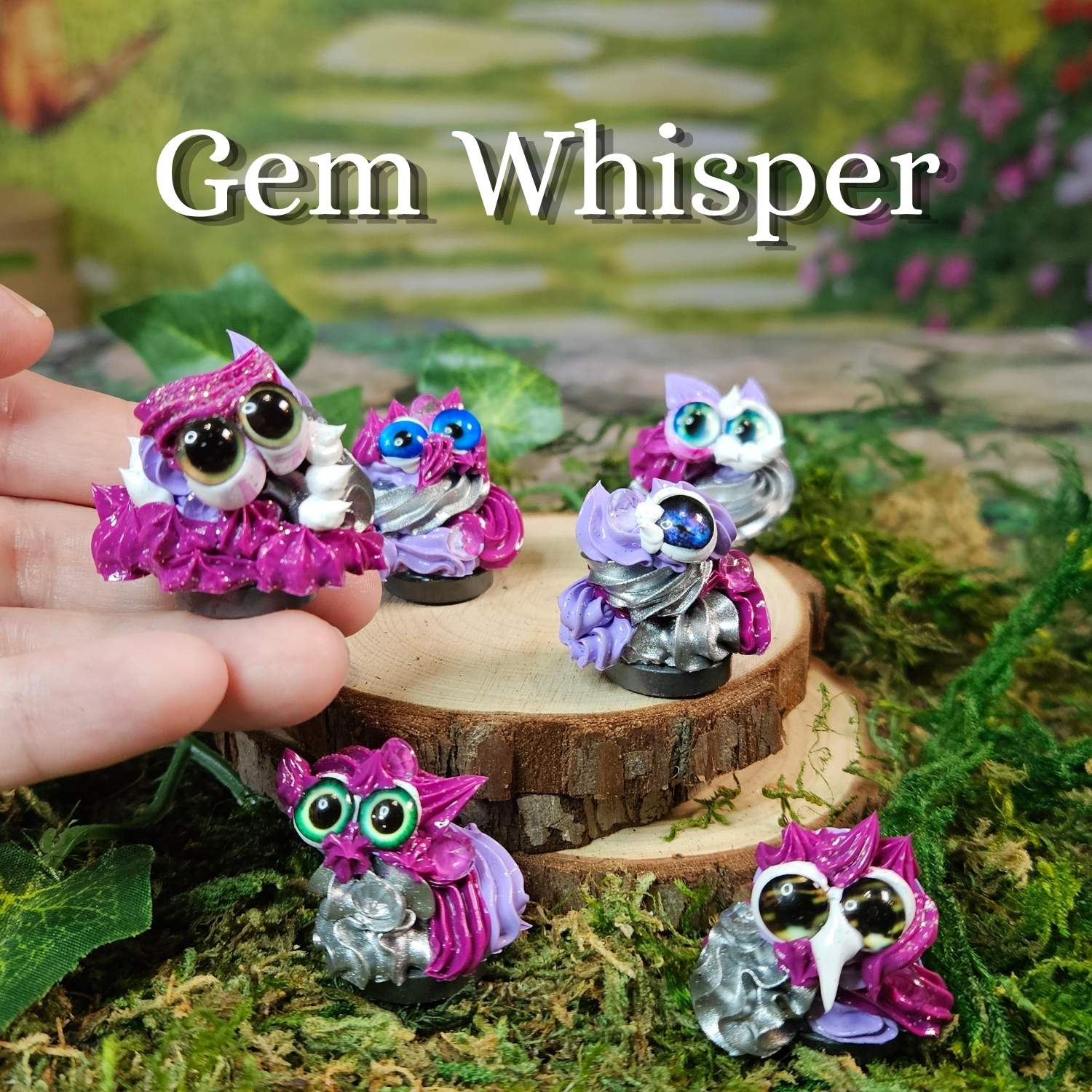 Five small frosting figurines with large glossy eyes and colorful glittery frosting texture are displayed on moss and a wood slice. One figurine is being held by a human hand, and the text 'Gem Whisper' appears at the top.
