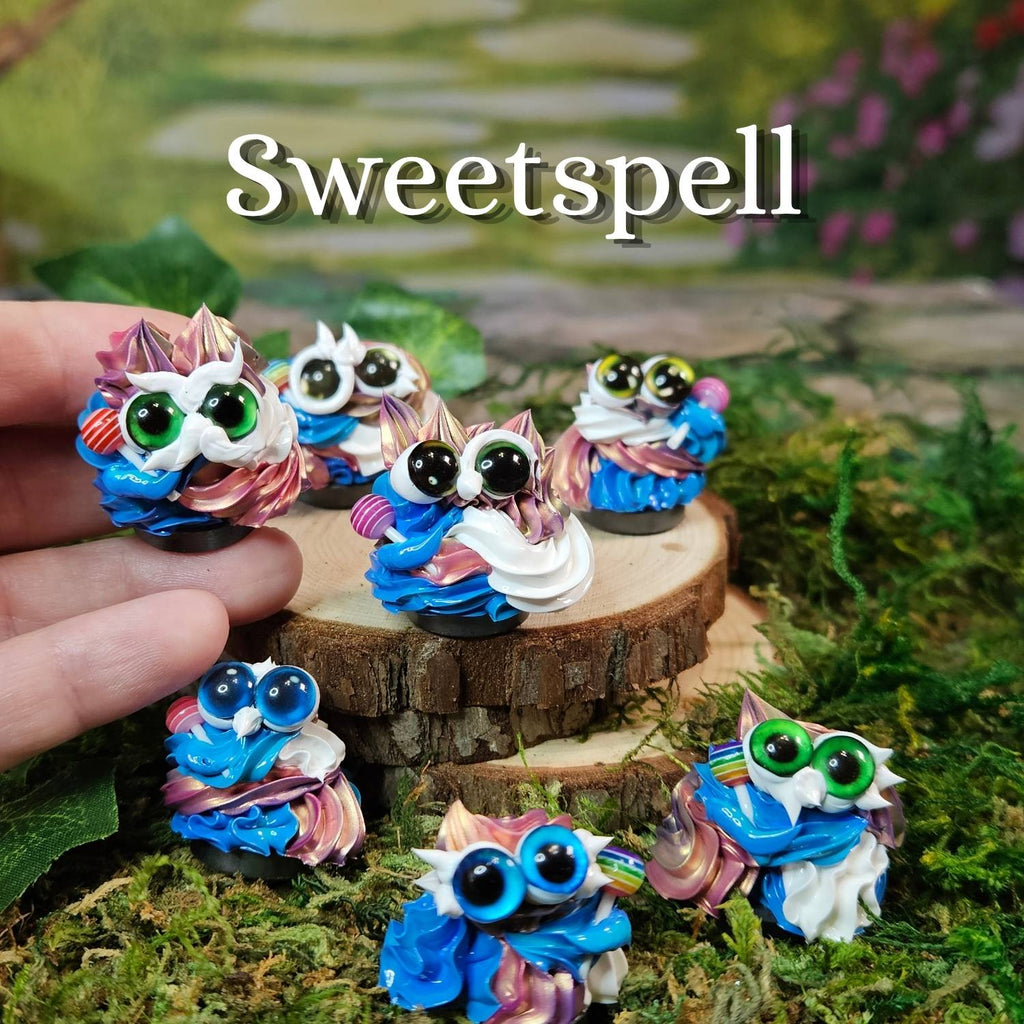 A hand holds a small colorful frosting figurine in front of several matching frosting decorations arranged on moss and a wood slice. The word 'Sweetspell' appears across the top.